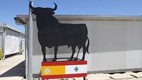 Die Silhouette eines schwarzen Stiers steht auf einem Podest in den Farben der spanischen Flagge, im Hintergrund ein Container