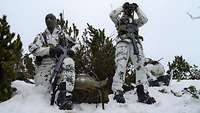 Ein Soldat steht im Schnee und beobachtet mit seinem Fernglas das Gelände. Neben ihm kniet ein weiterer Soldat mit Gewehr.