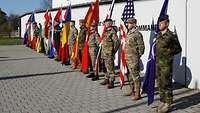 Soldaten verschiedener Nationen stehen mit ihrer Nationalflagge nebeneinander in einer Reihe vor einer Mauer