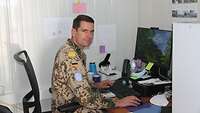 Ein Soldat sitzt in einem Büro vor seinem Computer und sieht in die Kamera
