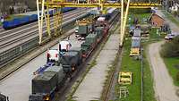 Ein Zug mit Containern und Militärfahrzeugen steht im Bahnhof unter einem Hebekran, der einen Container anhebt
