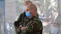 Ein niederländischer Soldat mit Mund-Nasenschutz hat die Arme verschränkt und hört einem Vortragenden zu.
