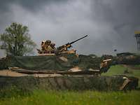 Ein Panzer mit Tarnnetzen und mit zwei Soldaten in Tarnfleckuniform im Turm auf einer grünen Wiese.