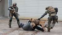 Ein Soldat hält einen Hund fest, der sich am Arm eines Zivilisten am Boden festgebissen hat.