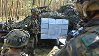 Ein Soldat steht im Wald vor einer Skizze, die an einem Kettenfahrzeug hängt, Soldaten hören ihm zu.