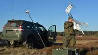 Ein Soldat steht auf einer Wiese vor einem Geländewagen mit einer Drohne.