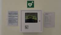 Ein Defibrillator in einem Kasten an der Wand; darüber ein Rettungszeichen: ein Herz mit Blitz darin und ein weißes Kreuz