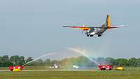 Zwei Feuerwehrautos stehen links und rechts neben einer Landebahn und verschießen Wasser auf ein landendes Flugzeug.