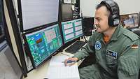 A man in an olive-colored overall with a headset on, sitting in front of several screens