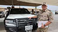 Ein Soldat mit einer Mappe in der Hand steht vor einem Geländewagen und lächelt in die Kamera