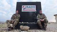 Zwei Soldaten sitzen von einem Schild mit der Aufschrift „Radio Andernach – Soldaten senden für Soldaten“