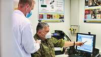 A soldier with a face mask explains something on the computer monitor to a man in a white gown