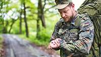 A soldier on a road in the woods. He is wearing a backpack and looks at his watch.