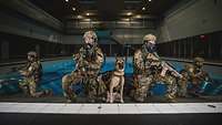 Vier bewaffnete Soldaten in Uniform und ein Hund hocken am Rand eines Schwimmbeckens.