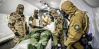 Mehrere Soldaten in ABC-Schutzkleidung stehen in einem Zelt um eine Krankentrage mit einer Person darauf