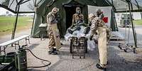 Vier Soldaten in ABC-Schutzkleidung stehen vor einem Zelt um eine Krankentrage mit einem Verletzten darauf