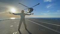 Ein Mann mit ausgebreiteten Armen schwenkt Sichtzeichen, ein Hubschrauber fliegt Richtung Flugdeck eines Schiffes