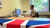 A British officer in uniform sits on his computer in his office.