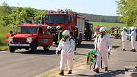 Rettungskräfte in weißen Vollkörperanzügen gehen in Richtung Hubschrauber, links auf der Straße stehen Feuerwehrwagen.