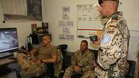 Zwei Soldaten sitzen in einem Büro. Im Vordergrund steht ein weiterer Soldat mit einer Kaffeetasse in der Hand