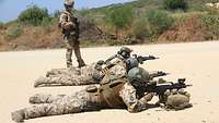 Drei Soldaten liegen nebeneinander auf dem Boden mit dem Gewehr im Anschlag. Ein weiterer Soldat steht hinter ihnen 