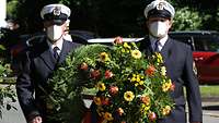 Zwei Marinesoldaten tragen einen Kranz, welchen mit Blumen geschmückt ist. Beide Soldaten Tragen eine Schutzmaske.