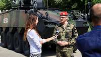 Eine Zivilistin mit Mikrofon steht mit einem Soldaten vor einem Panzer und interviewt ihn, ein Kameramann filmt die Szene. 