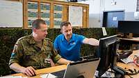 Ein Soldat und ein Mitarbeiter einer zivilen Firma sitzen zusammen an einem Computer. 