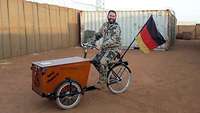 Soldat mit gelber Kordel sitzt auf einem Fahrrad welches vorn eine Kiste und am Gepäckträger eine Flaggenhalterung hat