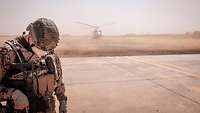 Ein Soldat mit gesenktem Kopf steht im Vordergrund. Er wendet sich von einem startenden Hubscharuber ab, der Sand aufwirbelt