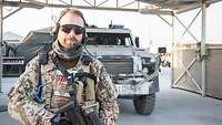 Ein Soldat mit Waffe, Sonnenschutzbrille und Gehörschutz mit Mikrofon steht vor einem Einsatzfahrzeug