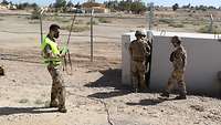 Ein Soldat mit Klemmbrett und Warnweste steht in der Wüste vor einem Schutzbau, den mehrere Soldaten betreten