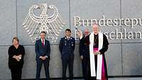 Eine Frau und drei Männer stehen vor dem Bundesadler. Davor segnet der Militärbischof mit Weihwasser die Flugbereitschaft.