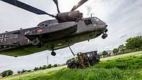 Ein Hubschrauber schwebt über einem Auto, daneben drei Soldaten, die das Auto mit einem Seil am Hubschrauber befestigen.