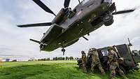 Ein Hubschrauber schwebt über einem Geländewagen. Neben dem Geländewagen stehen drei Soldaten.