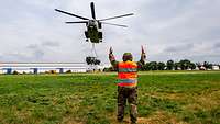 Ein Soldat steht auf einer Wiese. Vor ihm schwebt ein Hubschrauber mit einem Geländewagen.