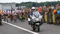 Eine Gruppe Radfahrer wird von uniformierten Menschen am Straßenrand bejubelt, im Vordergrund ein Motorrad.