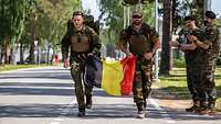 Zwei Soldaten laufen mit belgischer Flagge eine Straße entlang, rechts applaudieren zwei weitere Soldaten am Straßenrand