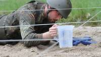 Ein Soldat schiebt einen Becher mit Flüssigkeit unter einem Gleithindernis hindurch.