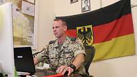 Ein Soldat sitzt an seinem Arbeitstisch vor zwei Monitoren. Hinter ihm an der Wand eine Deutschlandflagge