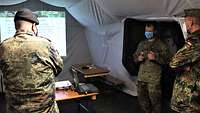 Drei Soldaten stehen in einem Zelt, im Hintergrund wird auf einer Leinwand eine Präsentation angezeigt.