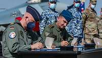 Zwei Soldaten sitzen an einem Tisch im Freien nebeneinander und unterschreiben jeder ein Papier