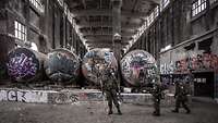 Drei Soldaten in ABC-Schutzkleidung in der Halle einer Industrieruine vor vier Tankbehältern mit Graffiti besprüht 