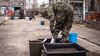 Ein Soldat in ABC-Schutzkleidung steht in einem Behälter mit Flüssigkeit und bürstet seine Schuhe