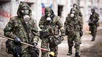 Fünf Soldaten in ABC-Schutzkleidung gehen hintereinander durch ein Fabrikgelände