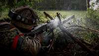 Ein Soldat schießt liegend in einem Waldstück mit einem Maschinengewehr.