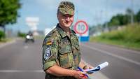 Ein soldat steht mit einem Klemmbrett in der Hand an einer Straße und lächelt in die Kamera