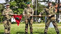 Drei Soldaten stehen nebeneinander auf einer Wiese und halten einen Wimpel mit Holzstange.