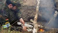 Ein Soldat kniet in einer Feuergrube vor einem Feuer und legt Holz nach