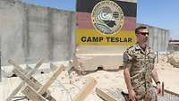 Ein Soldat steht vor einem militärischen Camp. Hinter ihm befindet sich ein Schutzwallelement mit der Aufschrift „Camp Teslar“